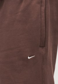 Rudi medvilniniai sportiniai kelnės su tekstūruota medžiaga, ant kairės pusės su baltu Nike logotipu ir šoninėmis kišenėmis funkcionalumui.