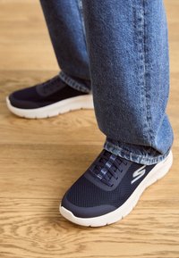 Marinblå nät-sneakers med vita sulor och texturerade detaljer, kombinerade med ljusblå denimjeans, visade på ett trägolv.