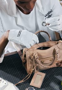 Personne portant des gants blancs inspecte un sac à main beige avec une loupe sur une surface de tapis de découpe noire.
