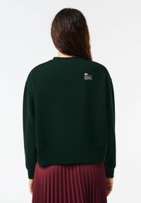 Zelená mikina vyrobená z bavlnenej zmesi, s okrúhlym výstrihom a rebrovanými manžetami. Na hornej časti chrbta je biele logo Lacoste.