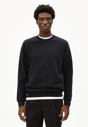 Schwarzer Sweatshirt aus weichem Material mit Rundhalsausschnitt und gerippten Bündchen, mit einem kleinen, tonalen Logo auf der Brust. Über einem weißen Hemd getragen.