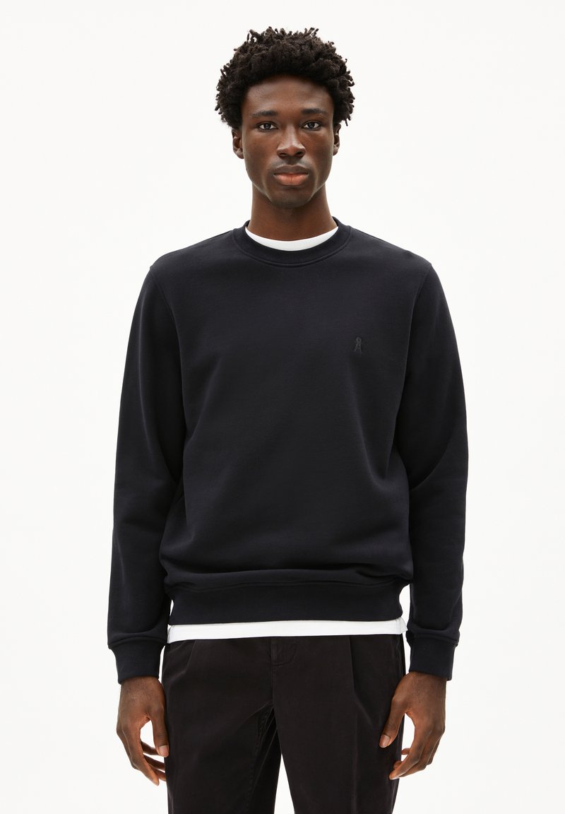 Felpa nera in tessuto morbido con collo rotondo e polsini a costine, con un piccolo logo tonale sul petto. Indossata sopra una camicia bianca.