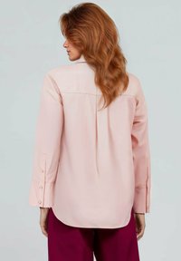 Lichtroze blouse met een comfortabele pasvorm, lange mouwen en knoopdetails op de manchetten. Heeft een rugplooi voor extra beweging en flow.