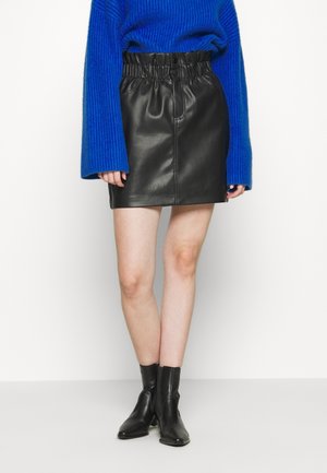 Person iført en sort læder miniskørt med rynket taljebånd, blå langærmet sweater og sorte ankelstøvler, stående mod en hvid baggrund.