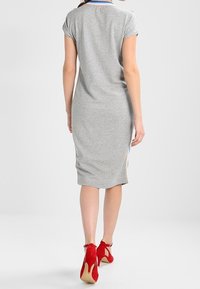 Kvinna som går bort ifrån kameran klädd i en grå knälång klänning med blå och beige sidostripes och röda högklackade skor på vit bakgrund.