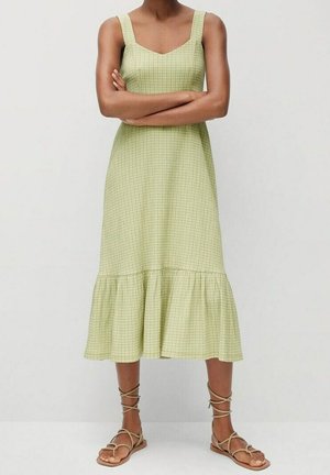 Grünes karierter Kleid mit quadratischem Ausschnitt, breiten Trägern und ausgestelltem Saum. Getragen mit sandfarbenen Schnürsandalen auf einem neutralen Hintergrund.