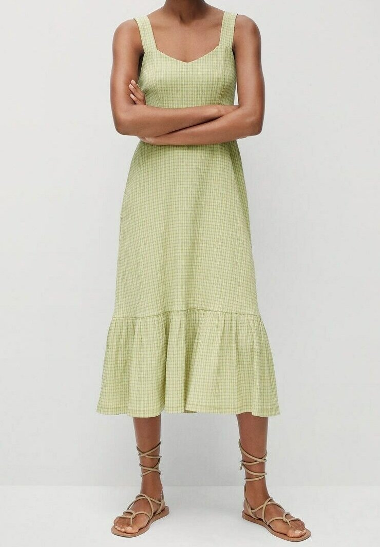 Robe en vichy vert avec un décolleté carré, des bretelles épaisses et un ourlet évasé. Portée avec des sandales à lacets beiges sur un fond neutre.