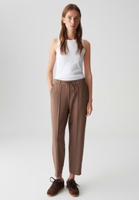 Jeune femme portant un haut sans manches blanc, un pantalon en tissu marron avec cordon de serrage et des baskets marron se tenant contre un fond clair.