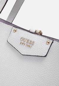 Hvid tekstureret lædertaske med guldtonet logo-patch og hardware, med sorte detaljer og en top lynlås-lukning.