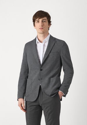 Blazer grigio a motivo con due tasche anteriori e risvolto a punta. Indossato sopra una camicia bianca, abbinato a pantaloni grigi.
