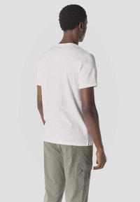Homme aux cheveux courts et bouclés, portant une chemise blanche à manches courtes et un pantalon cargo vert olive, debout face à un fond uni, de dos.