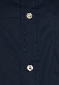 Camisa de gola azul escuro com dois botões grandes e redondos e um bolso de remendo. O tecido tem uma superfície lisa e texturizada.