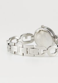 Silberne Metallarmbanduhr mit Gliederarmband, das "GUESS" und "G" Gravuren zeigt, von hinten auf weißem Hintergrund dargestellt.