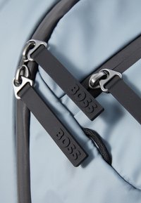Világoskék hátizsák, amely elegáns dizájnnal, texturált anyaggal és kontrasztos fekete cipzárahúzókkal rendelkezik, dombornyomott logótartozékokkal.
