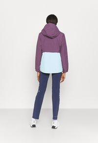 Chaqueta color bloqueada en púrpura y azul claro con capucha, que presenta un ajuste relajado. Combinada con pantalones azul marino y calzado blanco.