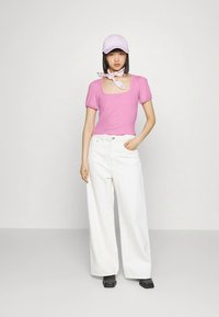 Top rosa canelado de manga curta com decote quadrado, combinado com calças largas brancas. Os acessórios incluem um boné lilás claro e um lenço estampado.