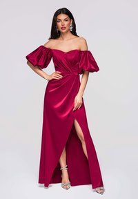 Robe en satin bordeaux avec manches bouffantes dénudées, corsage froncé et fente haute sur la cuisse. Accessoirisée avec des talons argentés à lanières et des bijoux.
