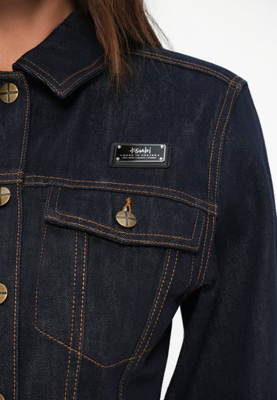 Σκουρό μπλε τζιν μπουφάν με χρυσές ραφές, μεταλλικά κουμπιά και προεξέχον ετικέτα επωνυμίας στην αριστερή τσέπη του στήθους.