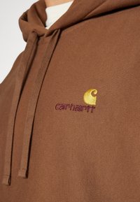 Felpa in cotone marrone con una finitura texturizzata. Presenta una tasca a marsupio e un logo ricamato giallo e viola sul petto.