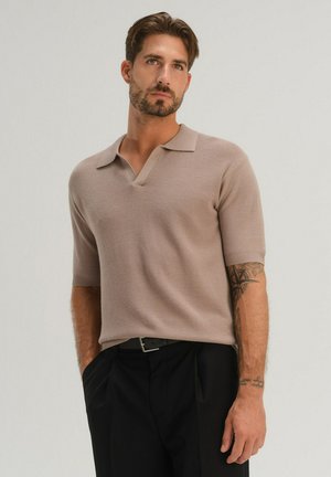 Man met baard en tatoeages, gekleed in een beige gebreide polo met korte mouwen en zwarte broek, staand met handen in de zakken tegen een neutrale achtergrond.