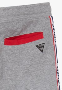 Grijze joggingbroek met rode zakrand, zwart-wit Guess-logo patch en een rode, witte en blauwe zijstreep met de tekst Guess.