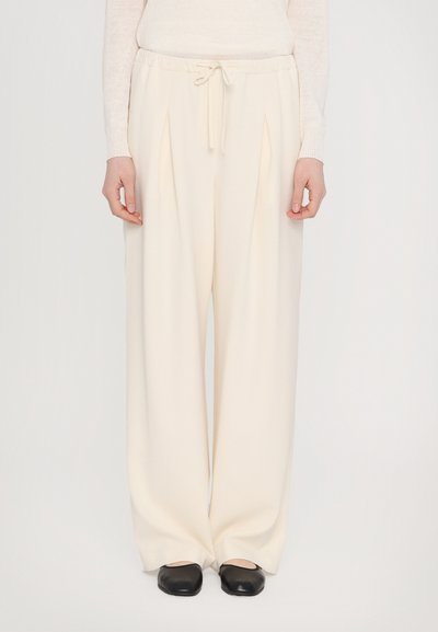 Pantalons beige à larges jambes avec une taille à cordon de serrage, présentant des plis à l'avant et une texture lisse. Associés à des chaussures noires à enfiler.