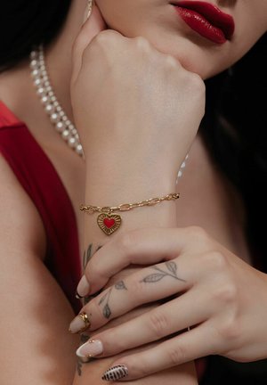 Gold chain bracelet with a red heart charm, featuring a textured design. Close-up of hands with nail art and fine line tattoos.