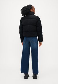 Svart puffjakke med høy krage, kombinert med vide blå jeans og svarte kraftige støvler. Teksturert stoff med synlige sømdetaljer.