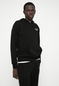 Sweatshirt preto com capuz e bolso frontal, material de algodão, design minimalista, com um logo no lado esquerdo do peito, combinado com calças a condizer.