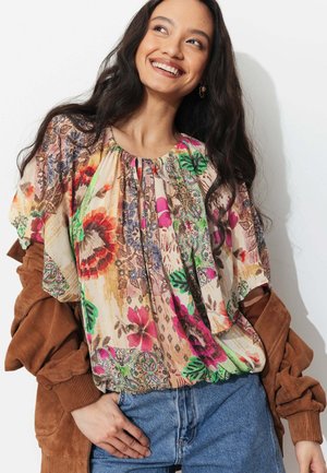 Femme souriante aux longs cheveux foncés portant un chemisier fleuri coloré sur une veste marron et un jean bleu, posant devant un fond blanc.