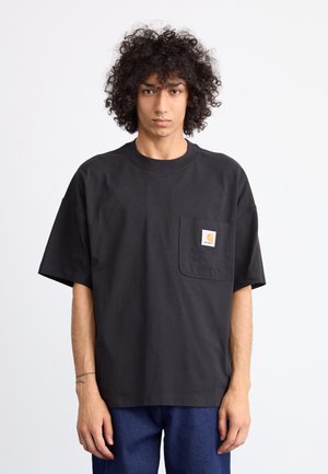 Young person with curly hair wearing a loose black t-shirt with a chest pocket and blue jeans, standing against a plain white background.