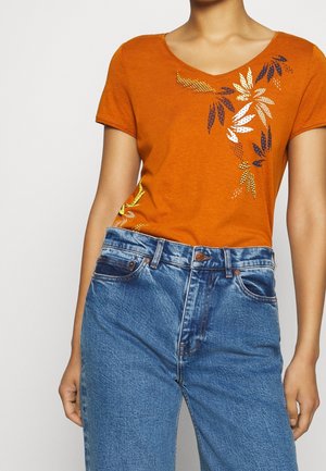 Nainen, joka pukeutunut oranssinväriseen lyhythihaiseen t-paitaan, jossa on kukkakuvio, ja korkeavyötäröisiin sinisiin farkkuihin yksinkertaista taustaa vasten.