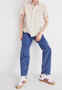 Krótki rękaw, beżowo-biała koszula w paski zestawiona z niebieskimi jeansami i białymi trampkami z czerwonymi i szarymi akcentami.