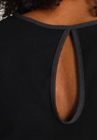 Schwarze Bluse mit einem Schlüsselloch-Ausschnitt auf der Rückseite, verziert mit einem kontrastierenden dunkelgrauen Besatz entlang des Halsausschnitts und der Kante des Ausschnitts.