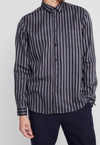 Camicia abbottonata con strisce verticali blu navy e grigie, maniche lunghe, tasca sul petto e colletto. Il tessuto sembra liscio.