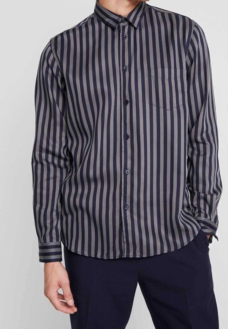 Camicia abbottonata con strisce verticali blu navy e grigie, maniche lunghe, tasca sul petto e colletto. Il tessuto sembra liscio.