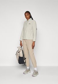 Giacca in pile grigia chiara con colletto alto, pantaloni beige, sneakers grigie, tiene uno zaino beige con base nera. Texture morbida, design funzionale.