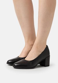 Chaussures à talons hauts en cuir noir avec un bout arrondi et un talon bloc épais, présentant une texture lisse et un design minimaliste.
