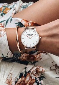 Orologio Victoria-Hyde in oro e rete con quadrante bianco e design minimalista, indossato con un bracciale in oro abbinato su una blusa a fantasia floreale.