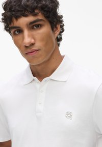 Fehér, rövid ujjú, pamut póló, három gombos nyakkal és egy kis ezüst logóval hímzéssel a mellrészen.