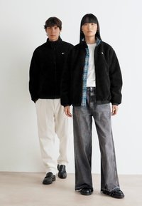 Two models stand against a white wall; one wears black fleece jacket and white pants, the other in black fleece jacket, plaid shirt, and gray jeans.