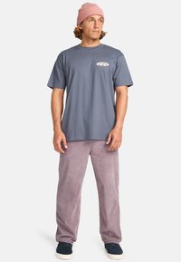 Camiseta de algodón gris con un pequeño logo en el pecho, combinada con pantalones de pana en un tono lila claro y un gorro de punto rosa.
