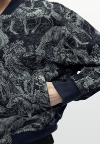 Κοντινή λήψη ενός σκούρου μπλε σακακιού με λεπτομερή γκρι σχέδια αλόγου και φύλλων, ριμπ μανσέτες και χέρι που έχει βάλει στην μπροστινή τσέπη.