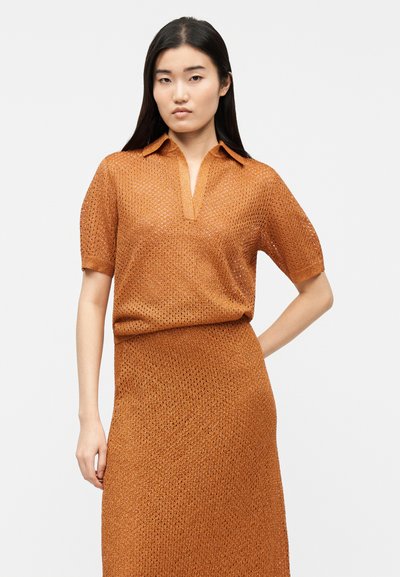 Νέα γυναίκα με μακριά σκούρα μαλλιά, φοράει έναν πορτοκαλί πλεκτό τοπ με κοντά μανίκια και φούστα που ταιριάζει, μπροστά σε απλό ανοιχτό φόντο.