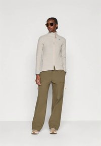 Beige-värinen vetoketjullinen takki, jossa korkea kaulus ja sininen merkki, yhdistetty löysiin oliivinvihreisiin housuihin. Malli käyttää aurinkolaseja ja neutraaleja tennareita.