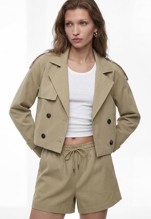 Kvinde iført beige dobbeltradet kort jakke og matchende shorts med hvid kort tanktop, stående med hænderne i lommerne.