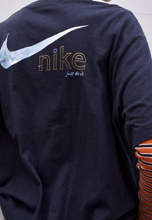 Osoba nosząca granatową koszulkę z dużym białym logo Nike w kształcie haczyka, złotym napisem „nike” oraz hasłem „just do it.” na plecach.