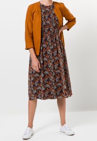 Chaqueta marrón de manga corta sobre un vestido floral con patrones en naranja, blanco y verde. Llevado con zapatillas blancas.