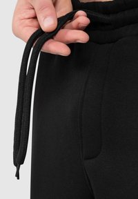 Sorte sweatpants med snøre i taljen, prydet med et tekstureret reb-snøre og sidelommer, vist med en hånd der holder snøret.