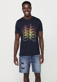 Camiseta de algodón azul marino con un diseño gráfico colorido de una bicicleta, de mangas cortas y cuello redondo. Combinada con unos pantalones cortos de denim desgastados.